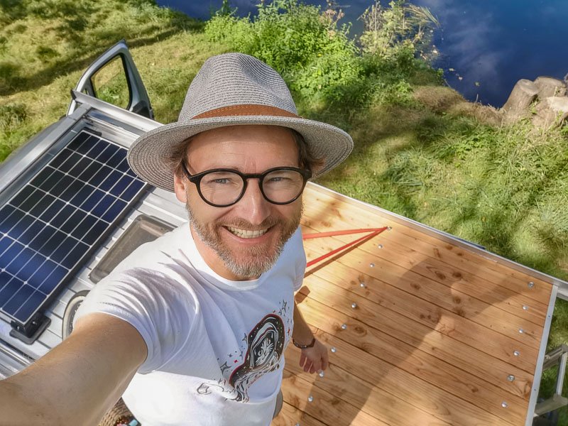 Steffen, Gründer von Arty Vans, mit Hut und ArtyVans Shirt auf der Dachterasse eines Campers an einem See.