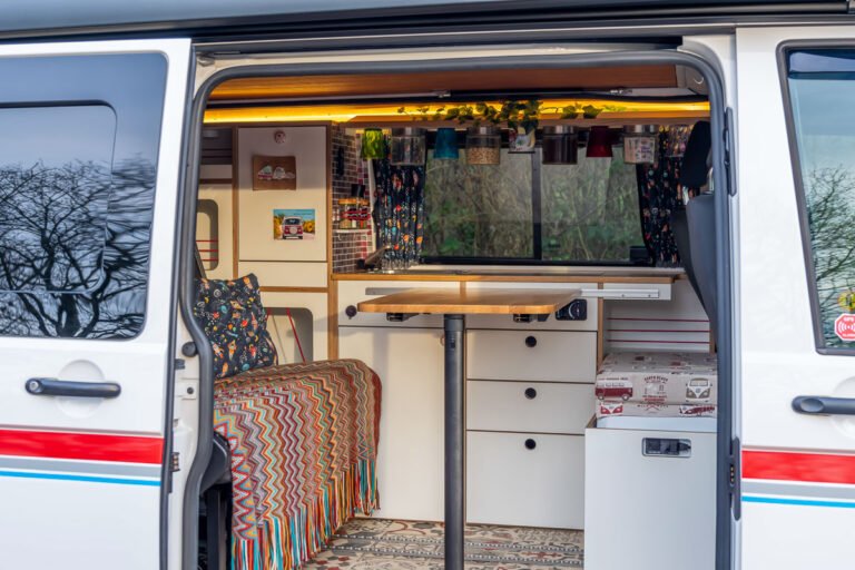 Projekt VW T6 37 VW T6 Arty Vans Camper Creation Innenraum. Blick von draußen durch die Schiebetür auf die Küchenzeile mit Eichenholz, Design-Vorhängen, Terrakotta Vintage Boden, VW Bulli Polster usw.
