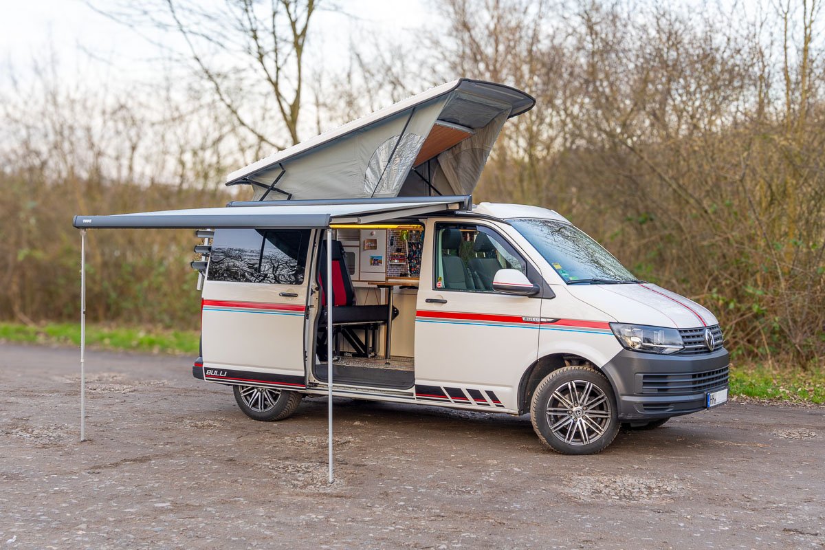 Arty Vans Creations VW T6 Bulli mit geöffnetem Hubdach und Markise auf einem Schotterweg in der Natur